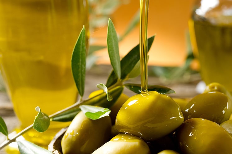 Olive oil being poured onto a bowl of green olives with an olive branch laying on the olives.