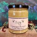 A jar of Champagne Roasted Garlic Mustard from The Olive Tap with a colorful background and surrounded by red and green decorative crinkle paper.