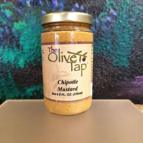 A 8 oz glass jar of Chipotle Mustard from The Olive Tap on a light colored surface twith a turquoise, black, and purple background.