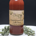 A glass bottle of Chipotle Roasted Garlic Grille Sauce from The Olive Tap surrounded by green decorative crinkle paper against a black background and white surface.
