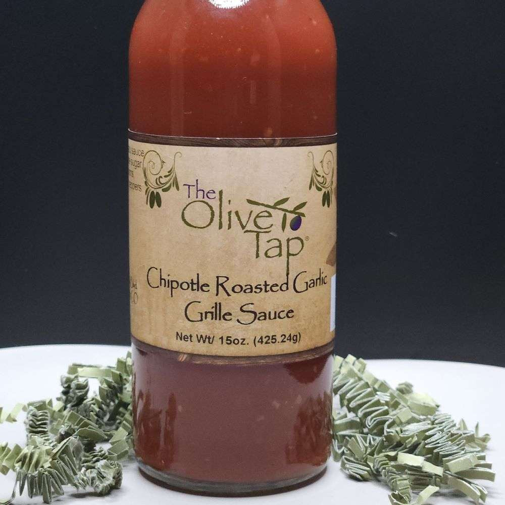 A glass bottle of Chipotle Roasted Garlic Grille Sauce from The Olive Tap surrounded by green decorative crinkle paper against a black background and white surface.