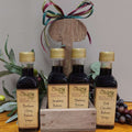 Cordial Sampler from The Olive Tap: four bottles of balsamic vinegars. Two of the bottles are in a wooden caddy, one more on each side. A bow on top of the caddy, and grapes and olives with leaves in the background.