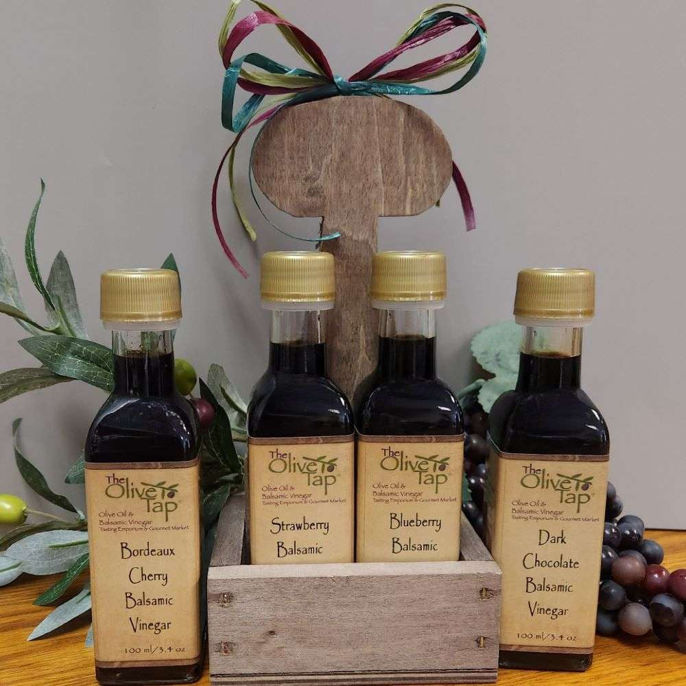 Cordial Sampler from The Olive Tap: four bottles of balsamic vinegars. Two of the bottles are in a wooden caddy, one more on each side. A bow on top of the caddy, and grapes and olives with leaves in the background.