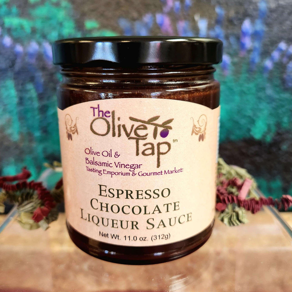 A jar of Espresso Chocolate Liqueur Sauce from The Olive Tap with a colorful background and surrounded by red and green decorative crinkle paper.