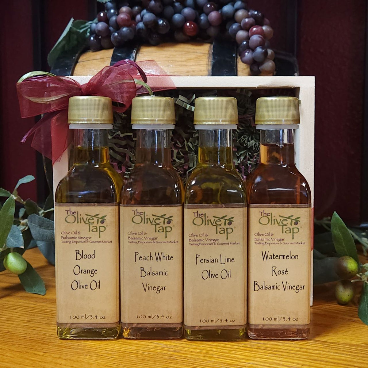 The Olive Tap's Fruity Sampler Summer Flavors: two olive oil bottles and two vinegar bottles in a wooden crate  with a bow surrounded by olive branches and grapes in the background.