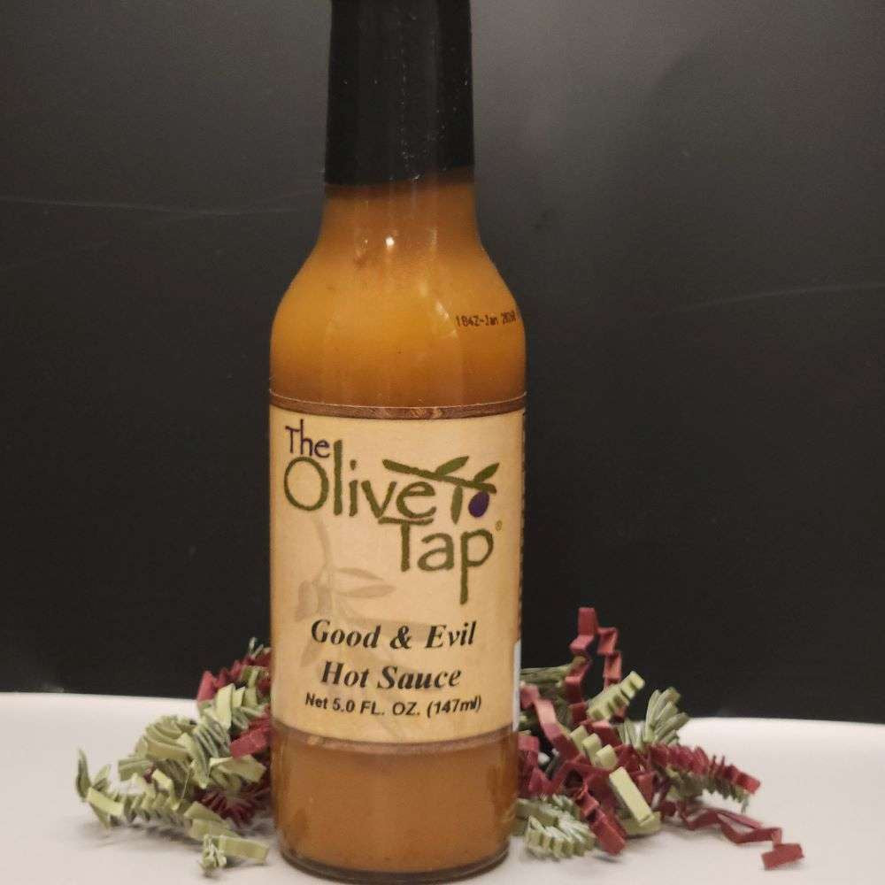A bottle of Good and Evil Hot Sauce from The Olive Tap surrounded by red and green decorative crinkle paper on a white surface against a black background.