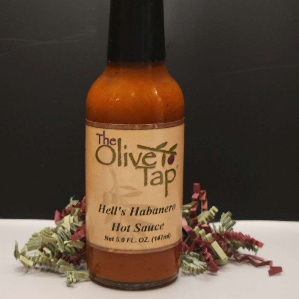 A glass bottle of Hell's Habanero Hot Sauce from The Olive Tap surrounded by red and green decorative crinkle paper on a white surface with a black background.