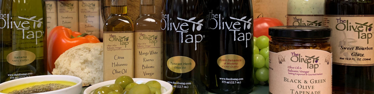 Collection of Olive Tap olive oil bottles, balsamic vinegar, and condiment jars surrounded by fruits and vegetables and bread with dipping oil.