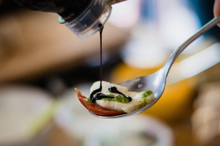 Spoon with a small dish of food being drizzled with a dark sauce on a blurred background.