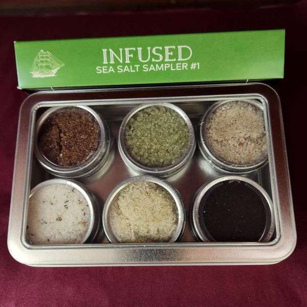 Infused Sea Salt Sampler Tin from The Olive Tap: Six round metal tins, each with their own infused sea salt inside, all within a rectangular metal tin and a green sign that reads "Infused sea salt sampler #1" against a red background.