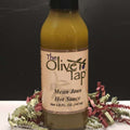 A glass bottle of Mean Jean Hot Sauce from The Olive Tap surrounded by red and green decorative crinkle paper on a white surface against a black background.
