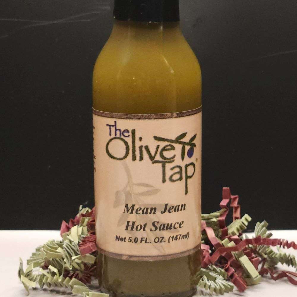A glass bottle of Mean Jean Hot Sauce from The Olive Tap surrounded by red and green decorative crinkle paper on a white surface against a black background.