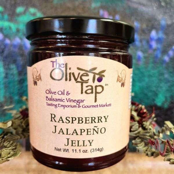 A jar of Raspberry Jalapeno Jelly from The Olive Tap with a colorful background and red and green decorative crinkle paper surrounding the jar.