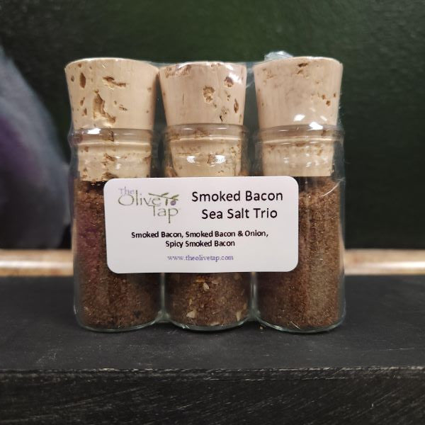 Smoked Bacon Sea Salt Trio from The Olive Tap: Three glass bottles of brown sea salts sealed with corks with a label on a dark surface on a green background.