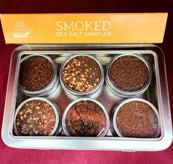 The Olive Tap's Smoked Sea Salt Sampler: six metal jars with brown flavored sea salts, two with yellow pieces mixed in, in a metal tray with a yellow sign that reads "smoked sea salt sampler".