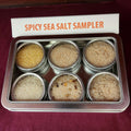Spicy Sea Salt Sampler Tin from The Olive Tap: An assortment of six spicy sea salts, each in their own metal tin, all within one larger rectangular tin with a sign that says "Spicy Sea Salt Sampler" against a red background.