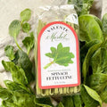 A package of Spinach Fettuccine from The Olive Tap on a light canvas background surrounded by fresh spinach.