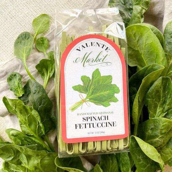 A package of Spinach Fettuccine from The Olive Tap on a light canvas background surrounded by fresh spinach.